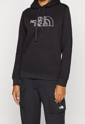 Osoba ubrana w czarną bluzę z kapturem The North Face z przednią kieszenią oraz dopasowane czarne spodnie bojówki z logo na prawej kieszeni.