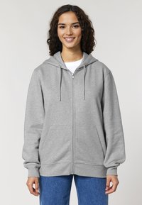 Felpa grigia con zip e cappuccio, tasche frontali e polsini a coste. Materiale morbido, vestibilità comoda, abbinata a jeans blu.