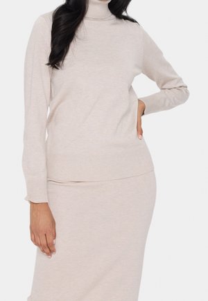 Kobieta ubrana w beżowy dzianinowy sweter z golfem i pasującą spódnicę, z ręką na biodrze, na jednolitym białym tle.