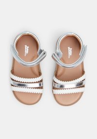 Srebrne metaliczne sandały z trzema białymi, ozdobionymi paskami, beżową podeszwą i regulowanym paskiem wokół kostki. Gładka faktura i płaski design.