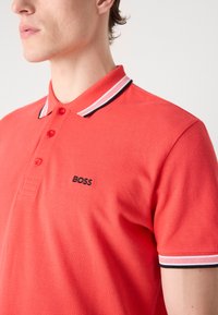 Rød polo-skjorte med teksturert stoff, med en to-knapps placket, brodert "BOSS" logo, og stripet kant på kragen og ermene.