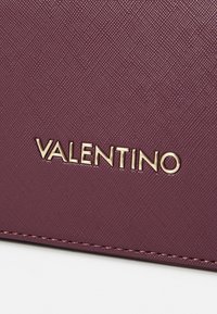 Sac en cuir violet avec une surface texturée, arborant le logo doré "VALENTINO". Comprend un détail de couture visible le long du bord.