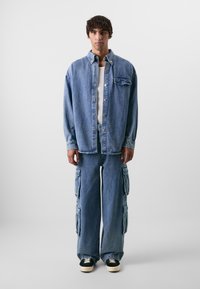 Camisa de ganga com um bolso frontal e botões de pressão, combinada com jeans cargo de perna larga com bolsos laterais. Ambos os itens são de um azul claro.