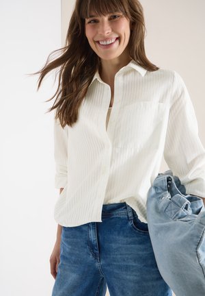 Smilende kvinde med brunt hår iført en hvid skjorte med knapper og blå jeans, der holder en lyseblå denimjakke.