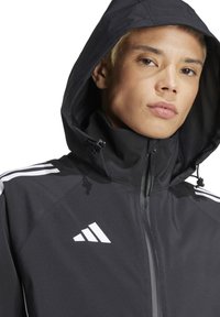 Schwarze sportliche Jacke mit Kapuze, mit einem Reißverschluss, weißen Drei-Streifen-Akzenten an den Schultern und einem Adidas-Logo auf der Brust.