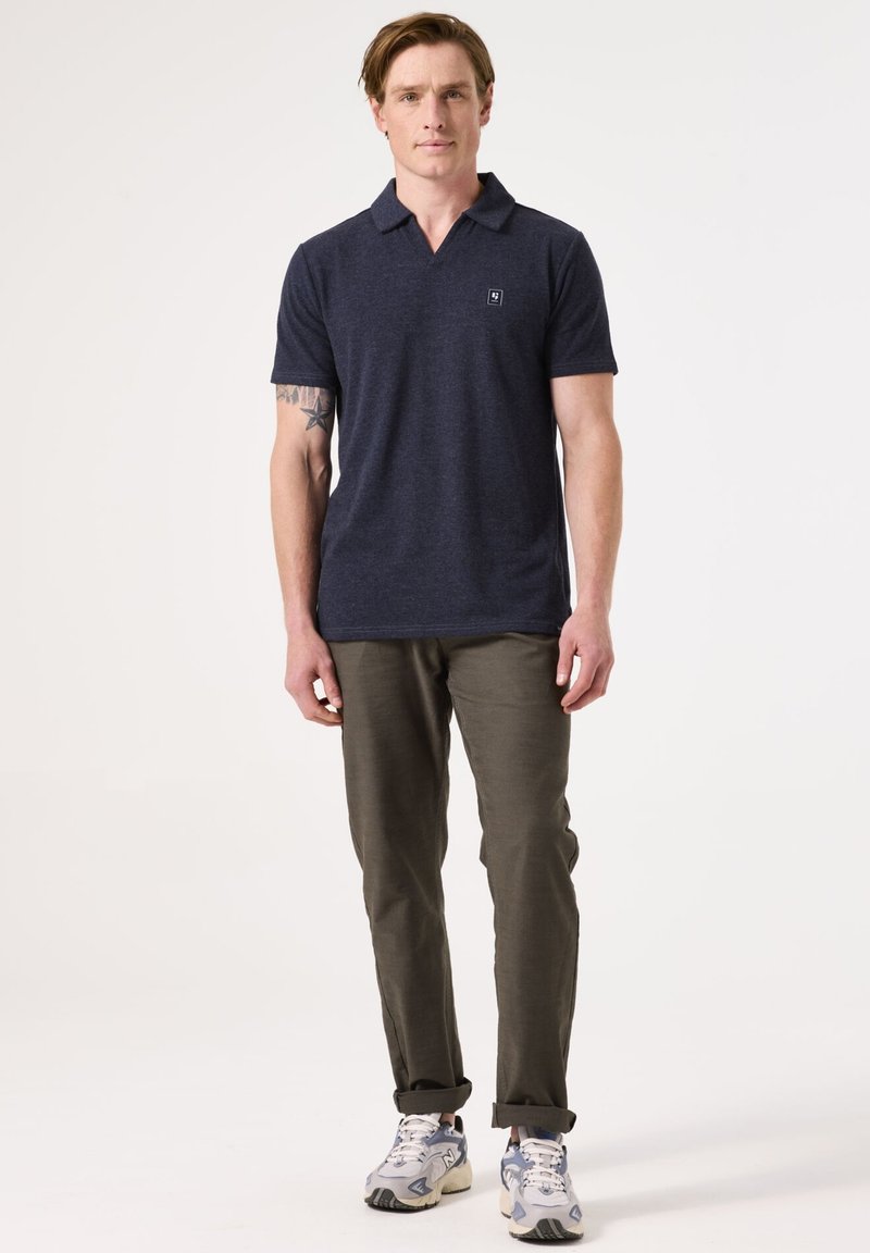 Marineblauwe poloshirt met een gestructureerde afwerking, korte mouwen en een subtiele logo. Gecombineerd met olijfgroene broek en grijze sneakers.