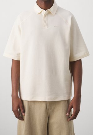 Homme portant un polo texturé à manches courtes couleur blanc cassé avec col et trois boutons, associé à un pantalon beige, visible du cou à la mi-cuisse.