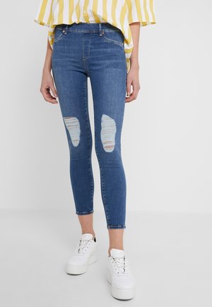 Personne portant un jean skinny bleu déchiré, des baskets blanches à plateforme et une chemise rayée jaune et blanche avec les manches retroussées.