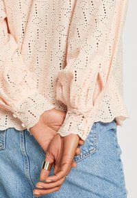 Mains croisées dans le dos, portant un chemisier rose pâle à œillets avec des poignets festonnés et un jean denim bleu clair, avec des bagues en or et petites.