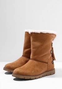 Mittelhohe braune Wildleder-Winterstiefel mit weißem Fellfutter, flachen Gummisohlen, Knopfdetails und Quasten an den Seiten.