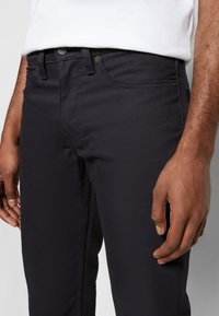 Närbild av en persons midja som visar svarta jeans och en vit skjorta, med en hand vila på sidan av jeansen.