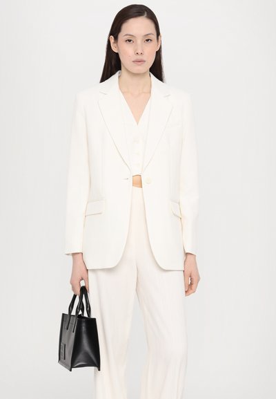 Blazer blanc ajusté avec des revers à cran, associé à un top blanc court et à des pantalons rayés. Le mannequin tient un petit sac à main en cuir noir.