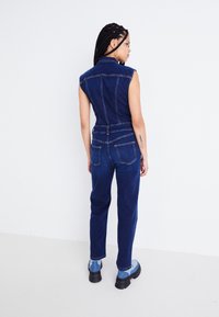 Combinaison en jean avec un corsage ajusté, des manches courtes et des pantalons à jambes droites. Tissu bleu foncé avec des coutures visibles et des poches arrière.