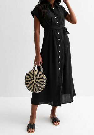 Femme en robe midi noire à boutons avec manches courtes, sandales noires sans lacets, tenant un sac à main rond tissé noir et blanc.