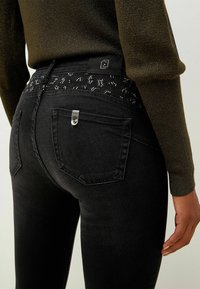 Vaqueros negros con un diseño ajustado, con una cinturilla texturizada adornada con un patrón de leopardo y acentos de hardware metálico.