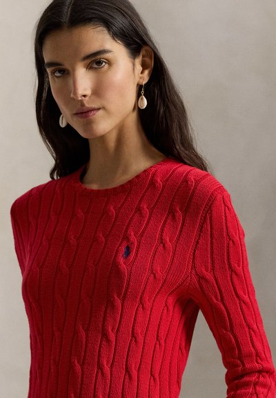 Γυναίκα με μακριά σκούρα μαλλιά, που φοράει λευκά σκουλαρίκια με κρεμαστό στοιχείο και κόκκινο πουλόβερ με ανάγλυφη πλέξη και μικρό σκούρο λογότυπο στο στήθος.