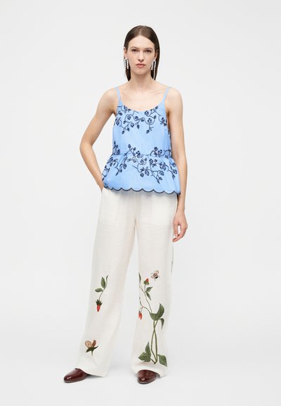 Γυναίκα που στέκεται με τα χέρια στις τσέπες, φοράει μπλε αμάνικο τοπ με κεντήματα λουλουδιών και λευκό παντελόνι φαρδιάς γραμμής με βοτανικά σχέδια.