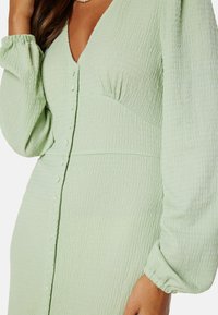 Abito verde chiaro in tessuto testurizzato con scollo a V, maniche lunghe a palloncino e chiusura con bottoni, caratterizzato da pieghe sottili e delicate arricciature.
