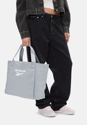 Osoba ubrana w czarne dżinsy i kurtkę, trzymająca jasnoszarą torbę Reebok, widoczne jasnoróżowe trampki.