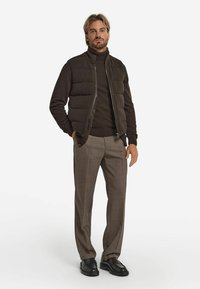 Dunkler Steppweste über einem braunen Rollkragenpullover, kombiniert mit hellbeigen karierten Stoffhosen und schwarzen Schuhen. Verfügt über eine gerippte Textur und einen Reißverschluss.