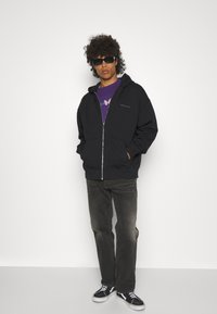 Czarna bluza z kapturem na zamek, z przednią kieszenią, noszona na fioletowej koszulce z grafiką, w połączeniu z szarymi spodniami i czarnymi butami do jazdy na deskorolce.