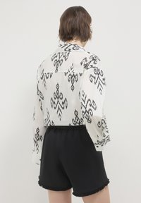 Blusa bianca trasparente con motivo tribale nero, maniche lunghe e colletto. Pantaloni corti neri con bordo sfilacciato, vita elastica e vestibilità rilassata.