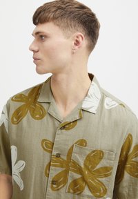 Camisa de manga corta, con botones, en verde oliva con grandes patrones florales en blanco y mostaza. Cuenta con un bolsillo en el pecho y un collar.