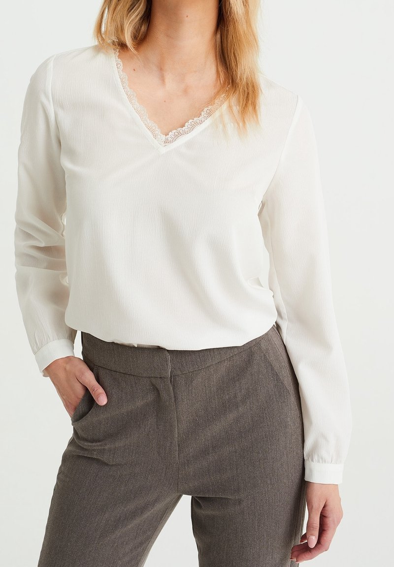 Femme portant un chemisier blanc à manches longues avec un col en V orné de dentelle et un pantalon gris taille haute, une main dans la poche, sur un fond clair.