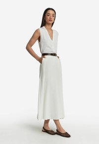 Weißes ärmelloses Oberteil mit V-Ausschnitt und Knöpfen, kombiniert mit einem schwingenden weißen Maxirock, der mit einem braunen strukturierten Gürtel und braunen Slingback-Schuhen gegürtet ist.