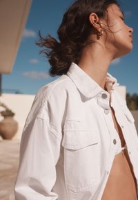 Giacca di denim bianca con collo a risvolto e due tasche sul petto. Caratterizzata da una vestibilità comoda e bottoni in metallo visibili. Sfondo di cielo azzurro e limpido.