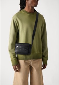 Felpa verde con polsini a coste, borsa a tracolla nera con logo in rilievo, indossata sopra pantaloni beige con vestibilità comoda.