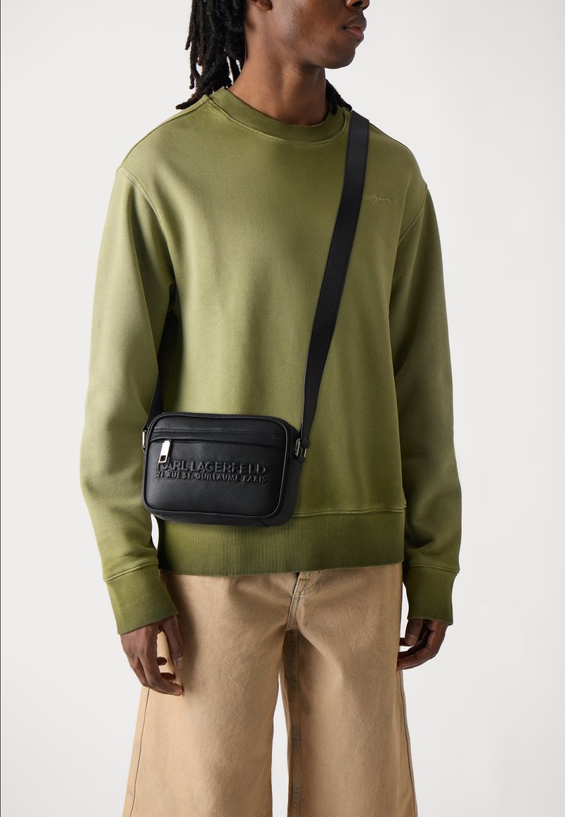 Felpa verde con polsini a coste, borsa a tracolla nera con logo in rilievo, indossata sopra pantaloni beige con vestibilità comoda.