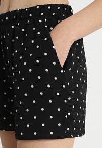 Femme portant un short noir à pois blancs, avec une taille élastique, et la main placée dans la poche droite.