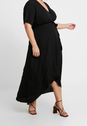 Kvinde iført sort omslagskjole med korte ærmer og bindebånd i taljen, iført brune, åbne højhælede sandaler, gående mod en enkel baggrund.