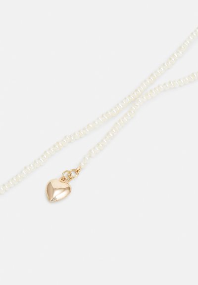 Duas tranças de pequenas pérolas brancas com um pingente de coração dourado polido preso por um fecho dourado. O coração é liso e refletivo.