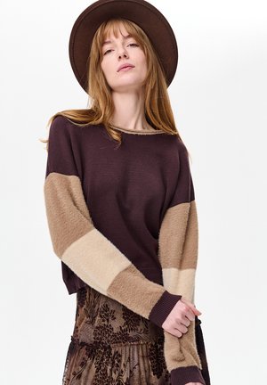 Bordeauxroter Strickpullover mit beigen, flauschigen Ärmeln, Rundhalsausschnitt, gerippten Bündchen, kombiniert mit einem gemusterten braunen Rock. Brauner breitkrempiger Hut.