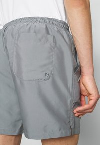 Personne portant un short gris avec une taille élastique et une poche arrière avec un petit détail circulaire dans le coin inférieur droit.