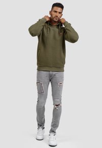 Sudadera con textura en verde oliva, con cordones y bolsillo frontal, combinada con jeans gris desgastados y zapatillas blancas.