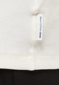 Λευκό ύφασμα με ραβδώσεις και κάθετη ετικέτα Marc O'Polo Denim ραμμένη κοντά σε ραφή πάνω από μαύρο υφασμάτινο φόντο.