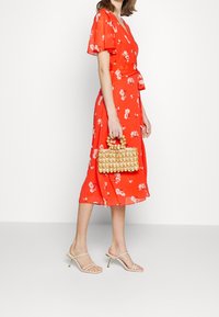 Robe fleurie rouge à manches courtes avec une taille nouée, associée à un sac à main beige à motif en treillis et des talons transparents à brides.