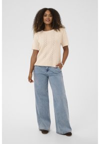 Ženska s kodrastimi lasmi, oblečena v bež pleteno majico s kratkimi rokavi, svetlo modre široke traperice, rjave čevlje in srebrno zapestnico, stoji na beli ozadju.