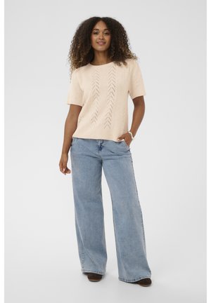 Kobieta z kręconymi włosami, ubrana w beżowy sweter z krótkim rękawem, jasnoniebieskie spodnie w stylu wide-leg, brązowe buty oraz srebrną bransoletkę, stojąca na białym tle.