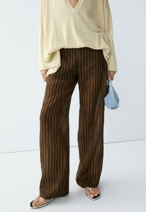 Personne portant un pantalon ample à rayures verticales marron et noir, un haut ample beige, des tongs blanches, tenant un petit sac à main bleu clair.