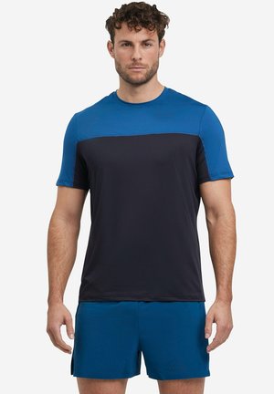 Mężczyzna w niebiesko-czarnym sportowym t-shircie z krótkim rękawem i pasujących niebieskich szortach, stojący zwrócony przodem na jednolitym tle.