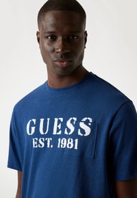 Tummansininen puuvillapaita, jossa on valkoiset painatukset "GUESS EST. 1981" ja pieni tasku. Pyöreä kaula-aukko ja lyhyet hihat.