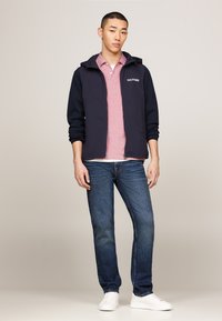 Giacca con cappuccio blu sopra una polo rossa, indossata con jeans blu e sneakers bianche. Design semplice, materiale in cotone e logo visibile sulla giacca.