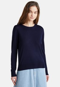 Mujer joven con cabello largo castaño, vistiendo un suéter de manga larga azul marino oscuro y una falda de mezclilla azul claro, contra un fondo blanco liso.