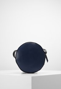 Kulatá tmavě modrá kožená crossbody taška s černým nastavitelným popruhem a zipem, vystavená na bílém podstavci na jednobarevném pozadí.