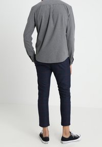Camicia grigia con chiusura a bottoni e orlo curvo, abbinata a pantaloni blu navy con motivo a griglia rossa. Sneakers nere completano l'outfit.
