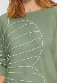 Jasnozielony sweter z białym haftem w kształcie fal, o luźnym kroju i miękkiej, teksturowanej tkaninie. Rękawy trzy czwarte dopełniają całość.